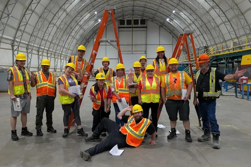 A group of IGVhousing workers in hard hats and safety vests posing inside the manufacturing facility