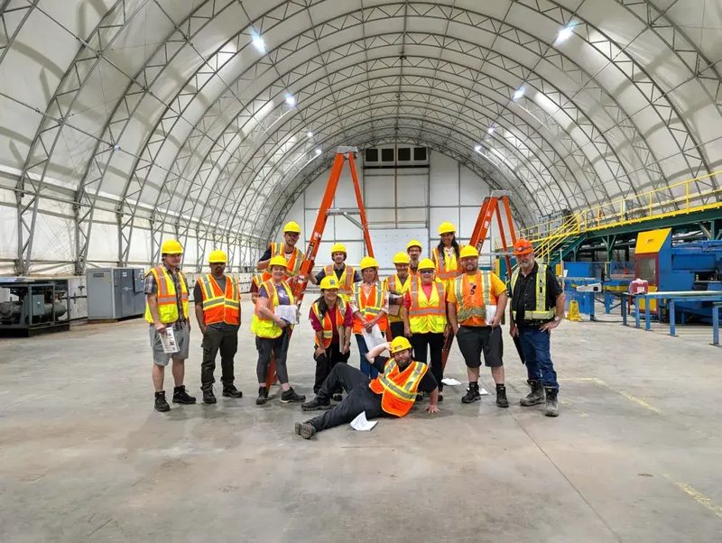 A group of IGVhousing workers in hard hats and safety vests posing inside the manufacturing facility
